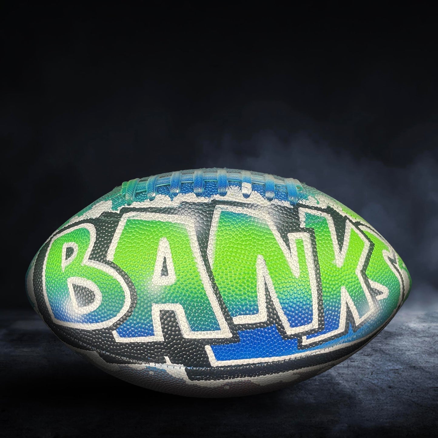 A football with the word "BANKS" painted on it, sitting on a dark surface with smoke or fog in the background.