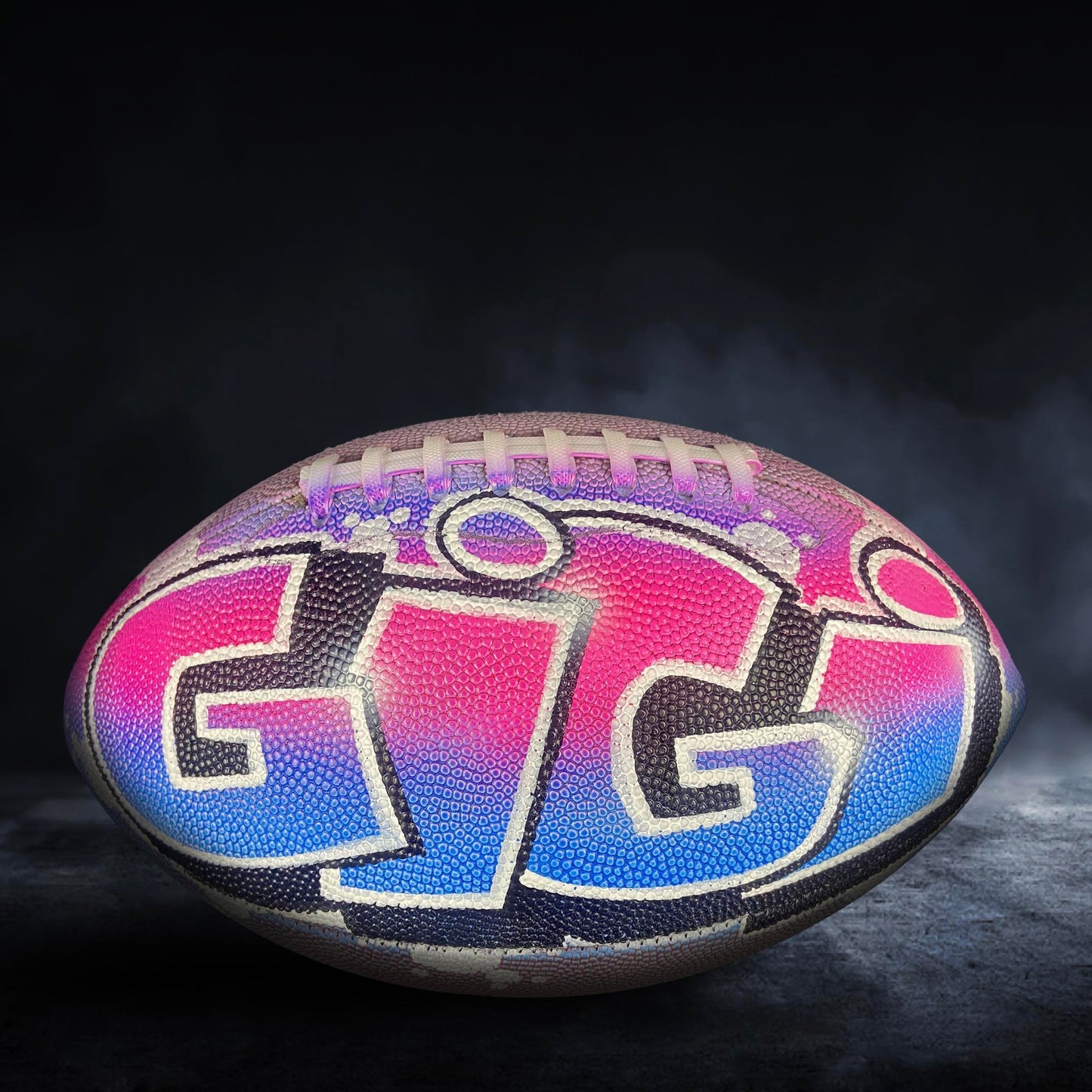 A colorful football with the letters "GIG" painted on it, sitting on a dark surface with smoke or fog in the background.