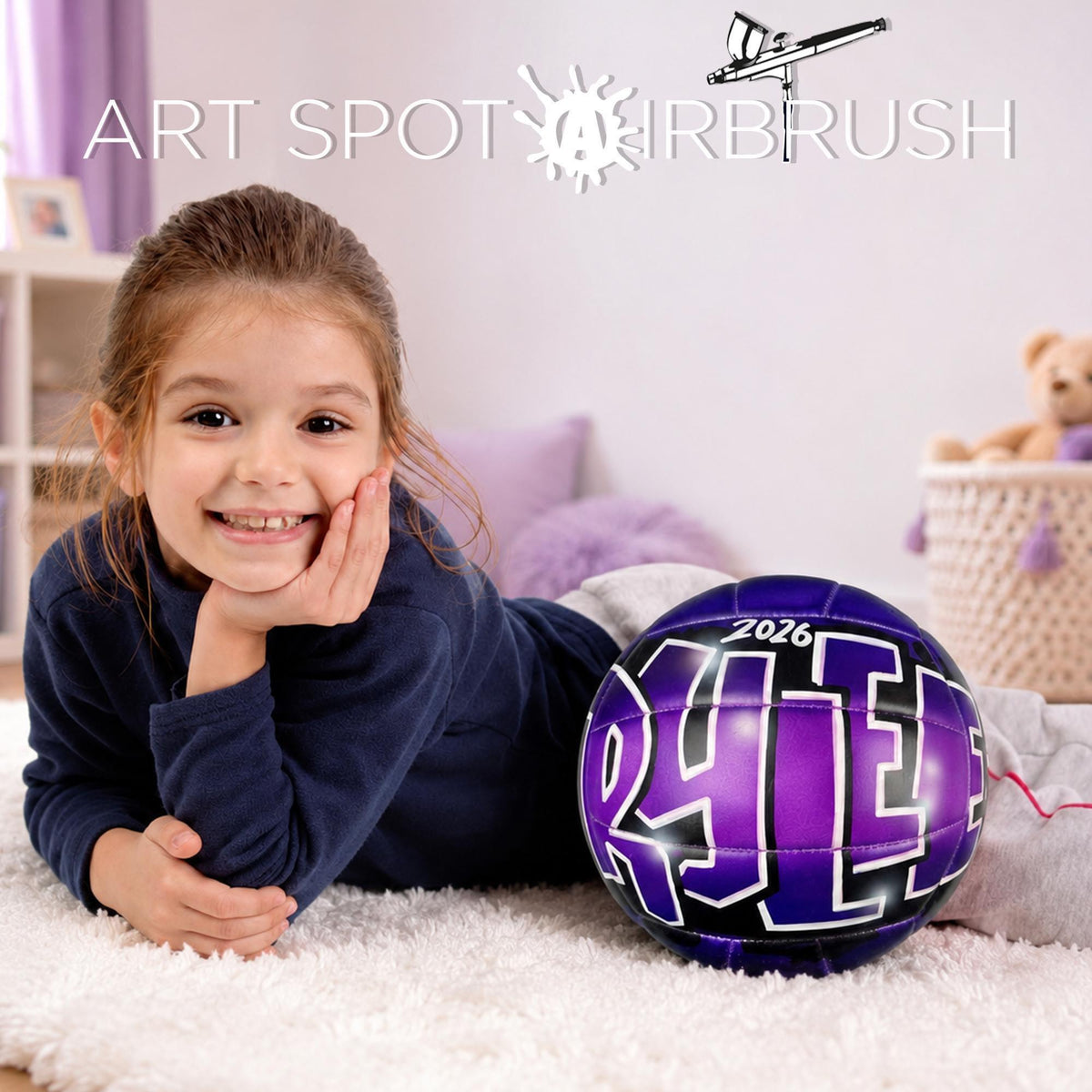 A young girl in a navy blue shirt is smiling and posing next to a purple custom volleyball with the number "2026" on it.