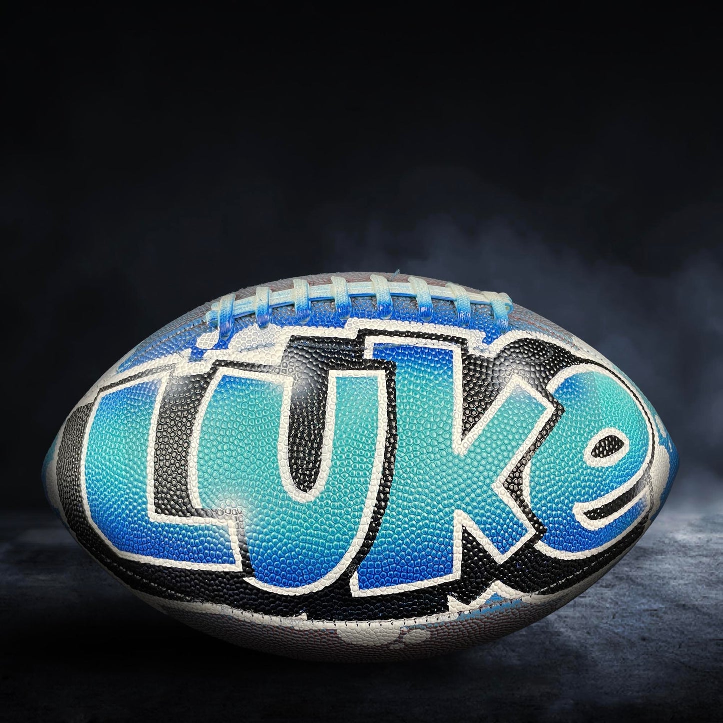 A football with the name "LUKE" painted on it, set against a dark background with smoke or fog.