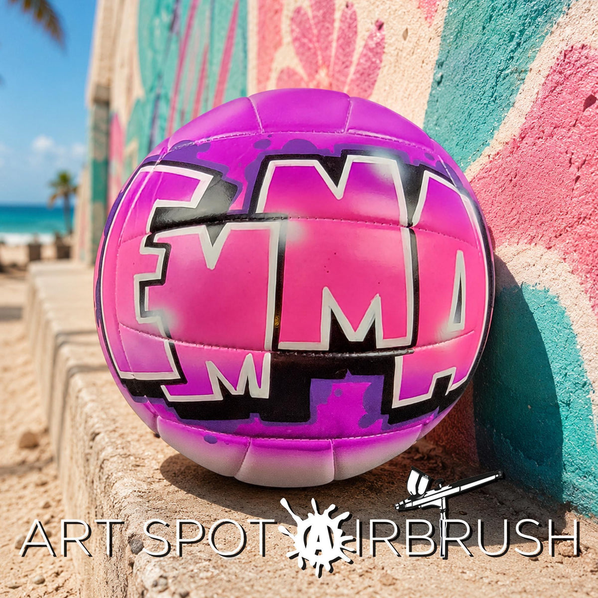 A pink and purple custom volleyball with the word "EMMA" painted on it, sitting on a beach with a colorful mural in the background.