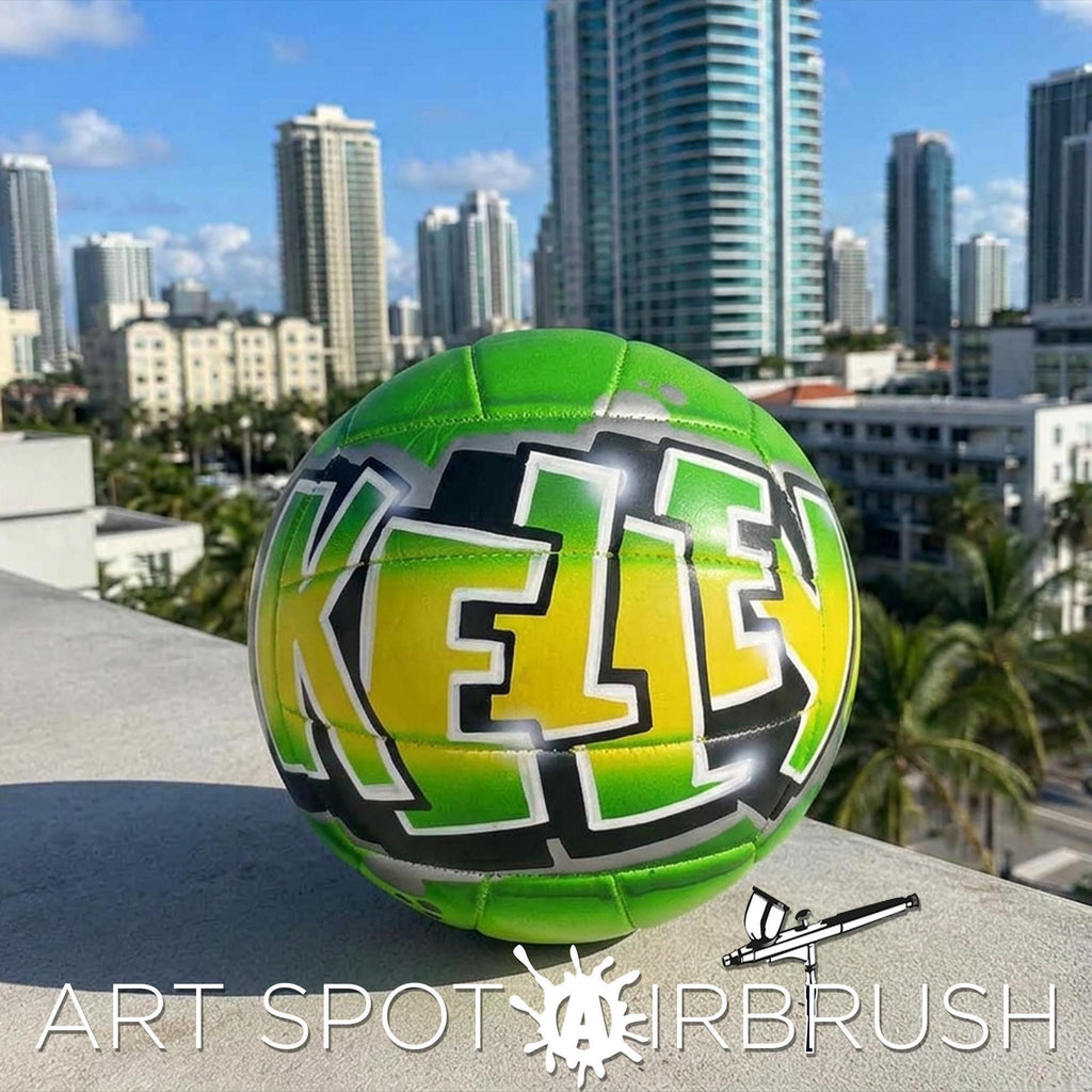 A green and yellow personalized custom volleyball with the name "KELLEY" painted on it, set against a backdrop of a city skyline.