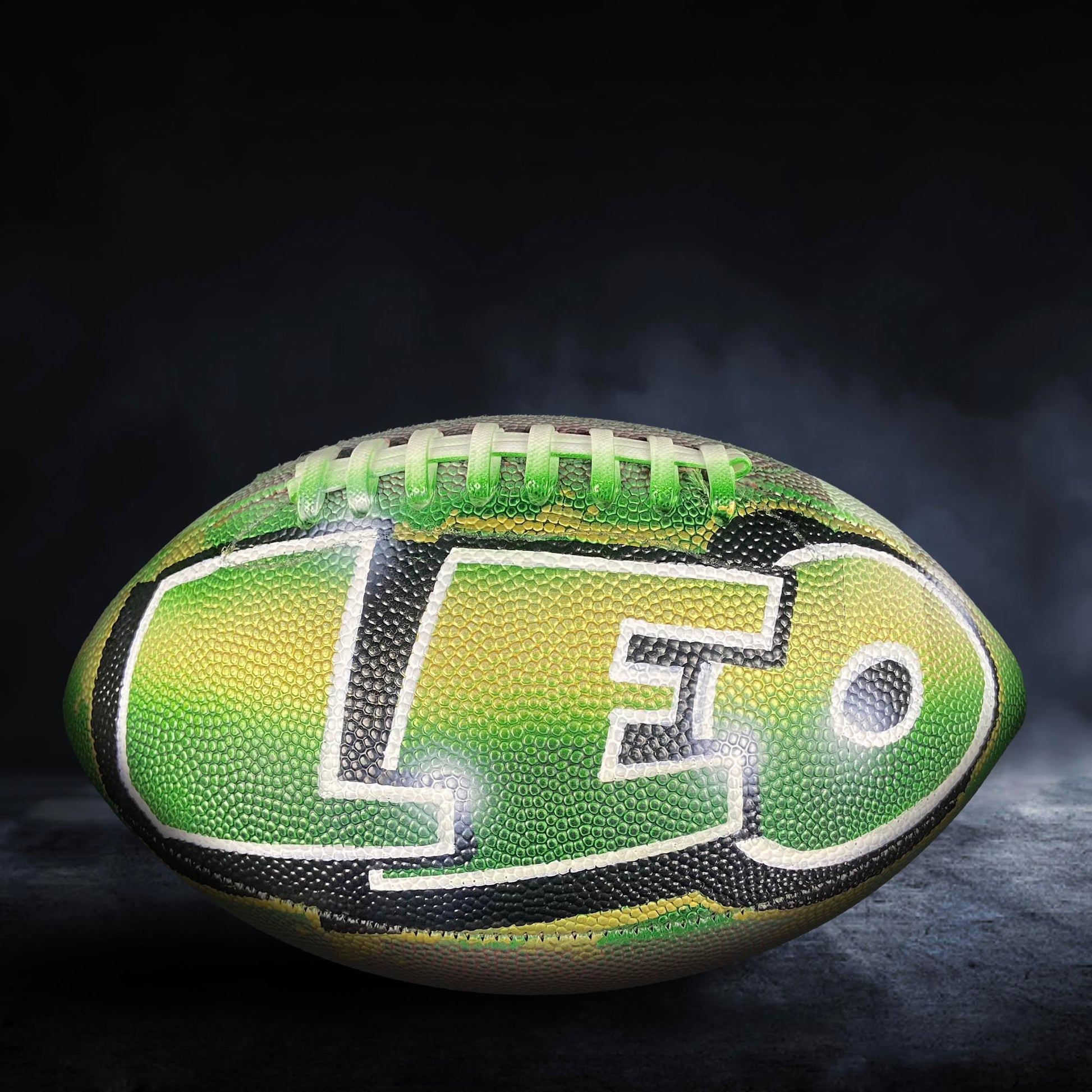 A green and black football with the word "LEO" written on it, placed on a dark surface with smoke or fog in the background.