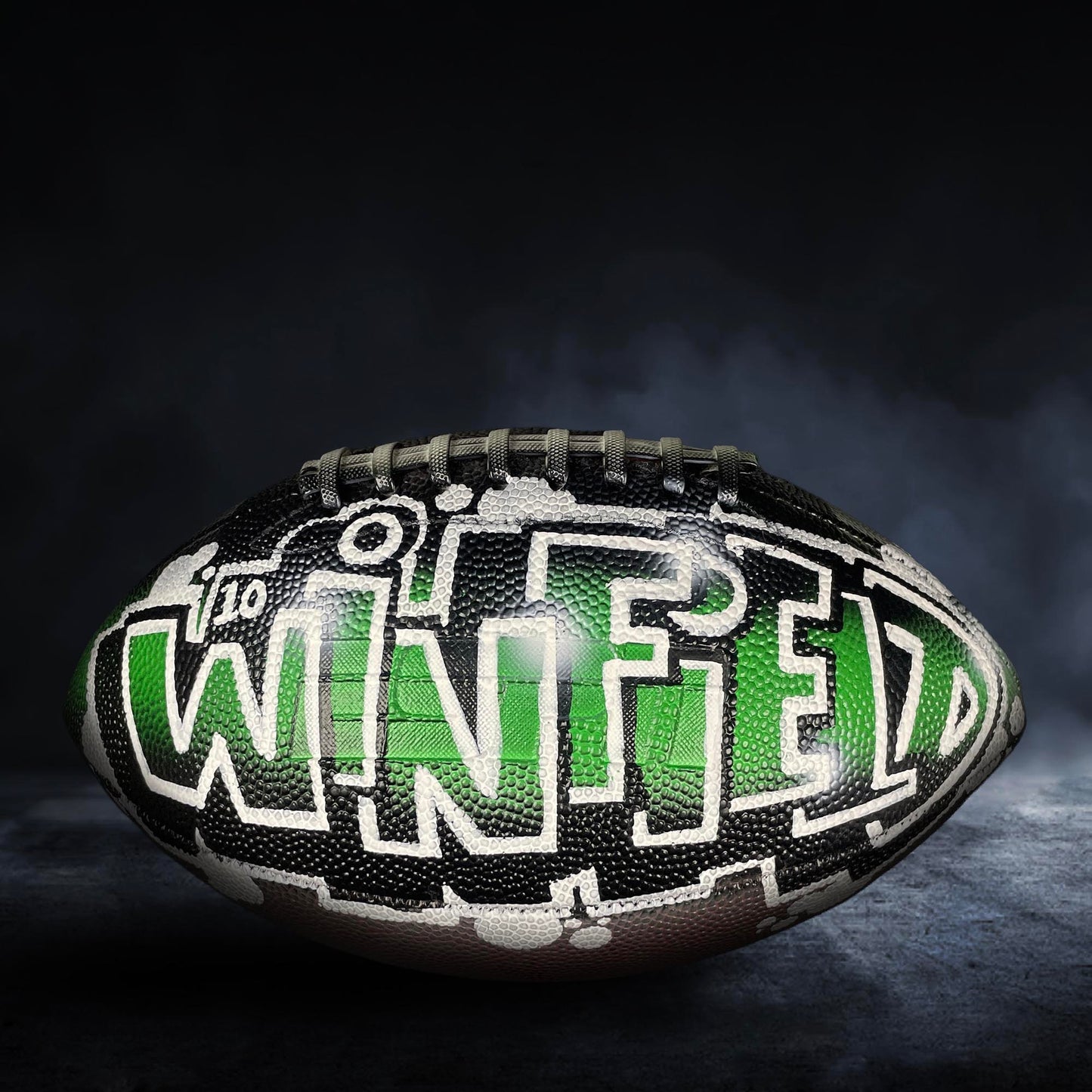 A football with the word "WINFIELD" painted on it, set against a dark background with smoke or fog.