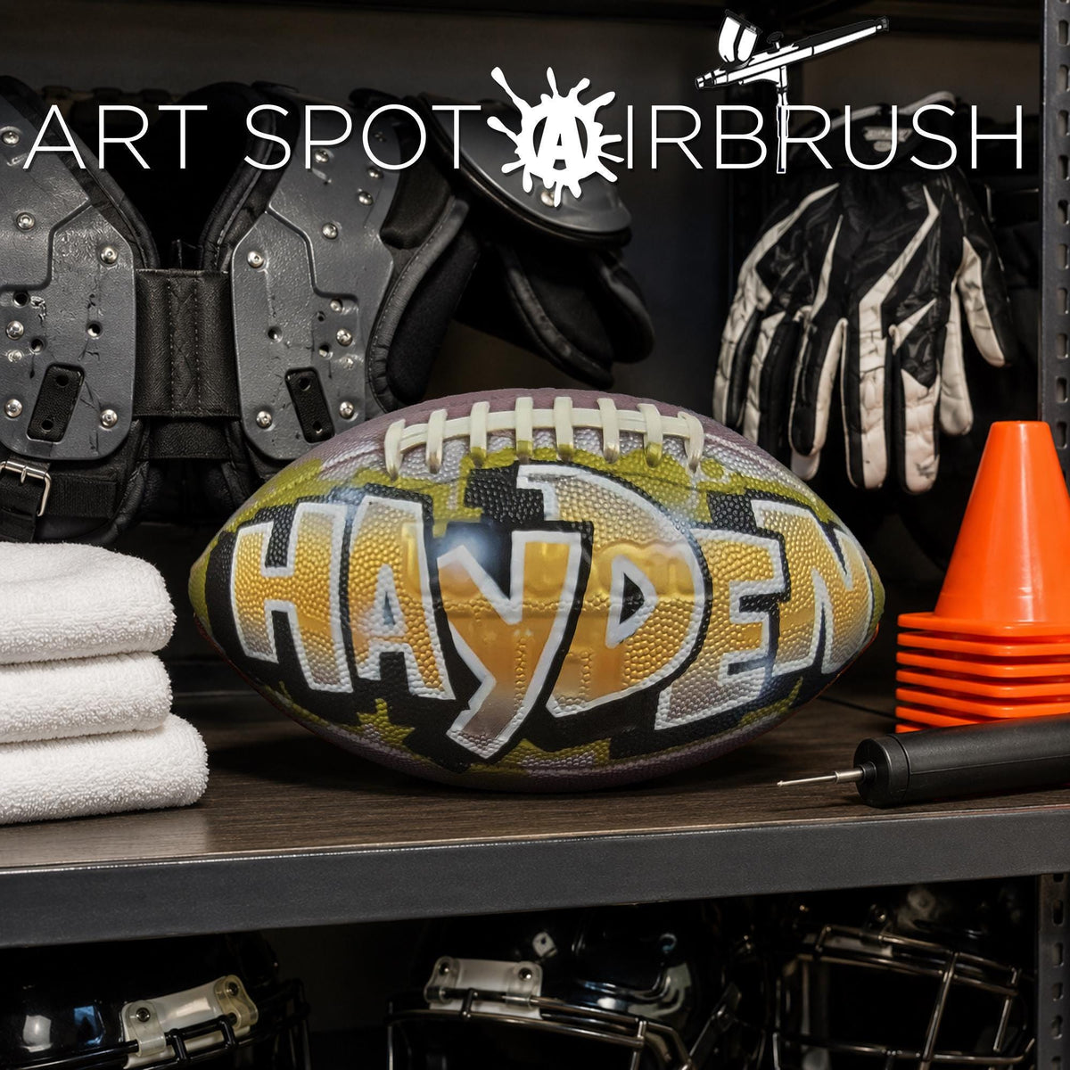 A custom football personalized with the name "HAYDEN" painted on it, along with various sports equipment such as helmets, pads, and cones, all displayed on a shelf.