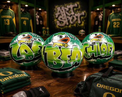 three green soccer balls with the word "BEA" written on them, placed on a wooden surface. In the background, there are several green jerseys and a sign that says "ART SHOT".