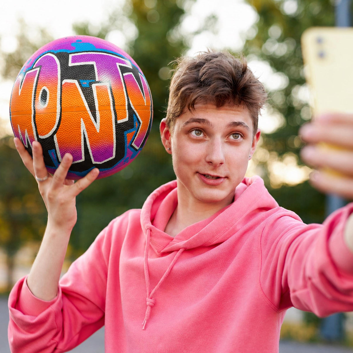 A young man in a pink hoodie is holding a colorful basketball with the word "MONTY" written on it, taking a selfie with a smartphone.