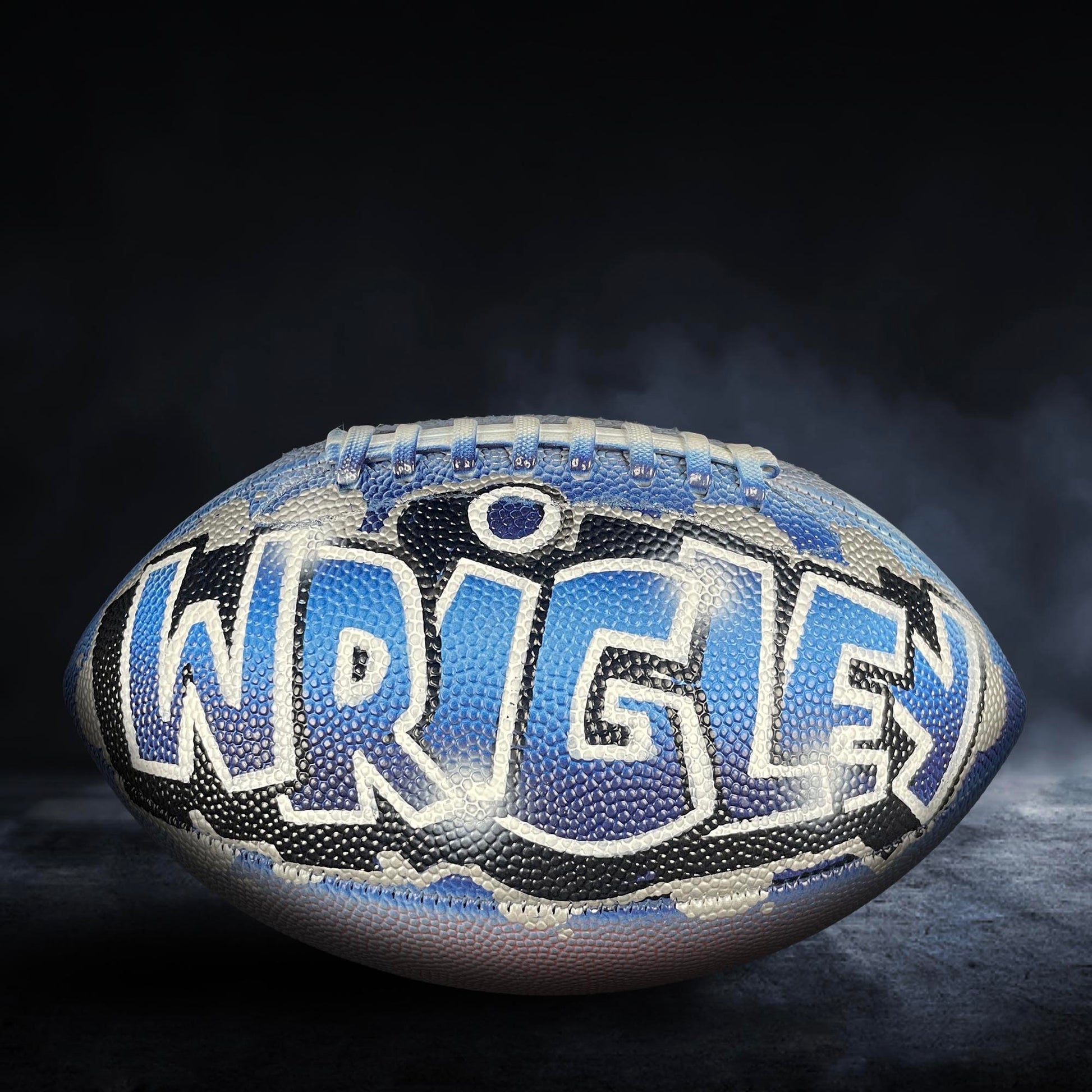 A blue and black football with the name "WRIGLEY" written on it, sitting on a dark surface with smoke or fog in the background.