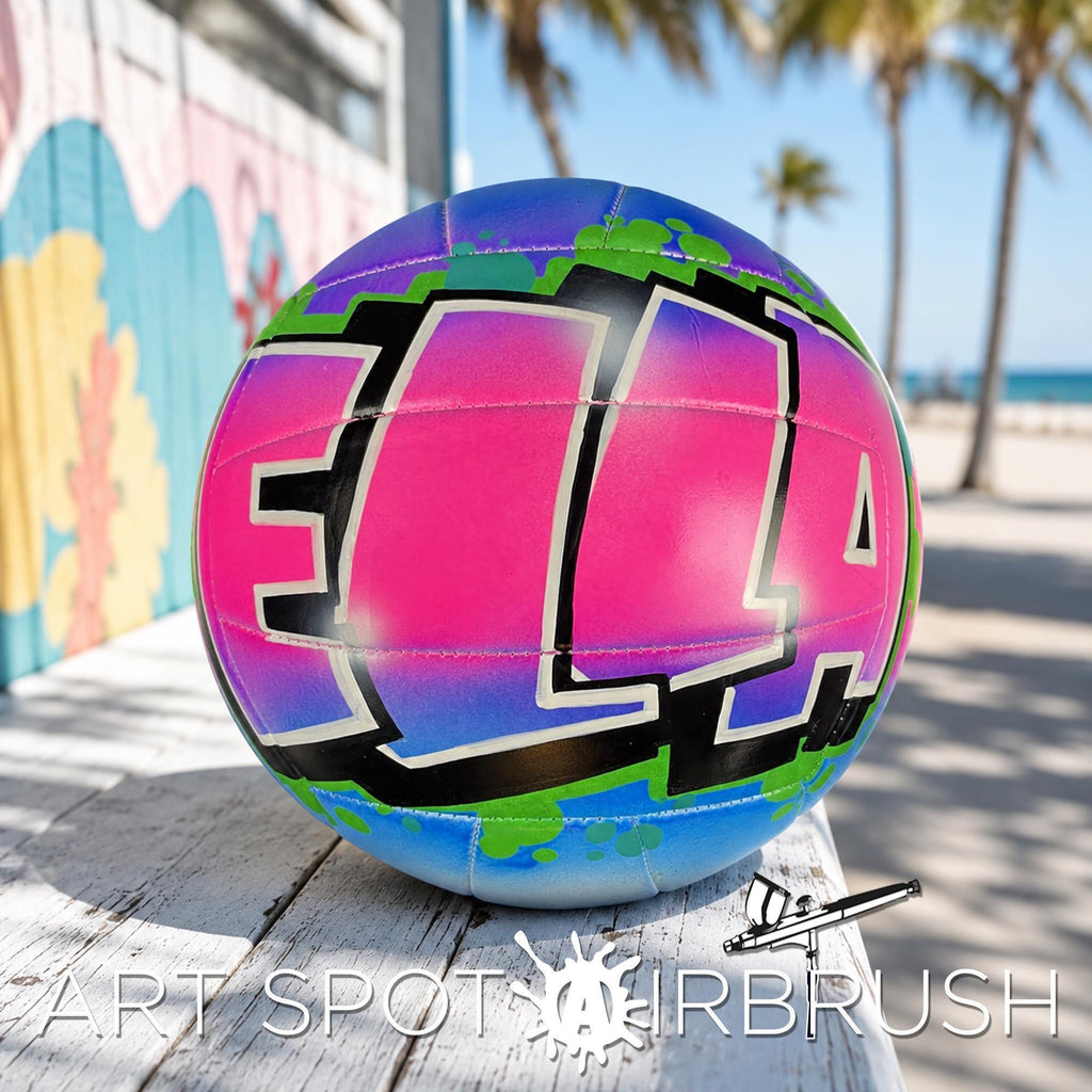 A colorful, graffiti-style custom volleyball personalized with the word "ELLA" painted on it, sitting on a wooden surface with palm trees and a beach in the background.