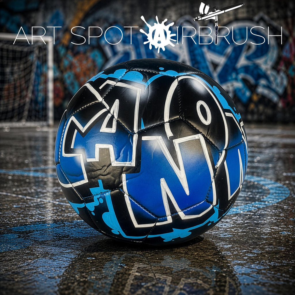 A soccer ball with graffiti-style lettering is displayed on a wet surface, with a graffiti-covered wall in the background.
