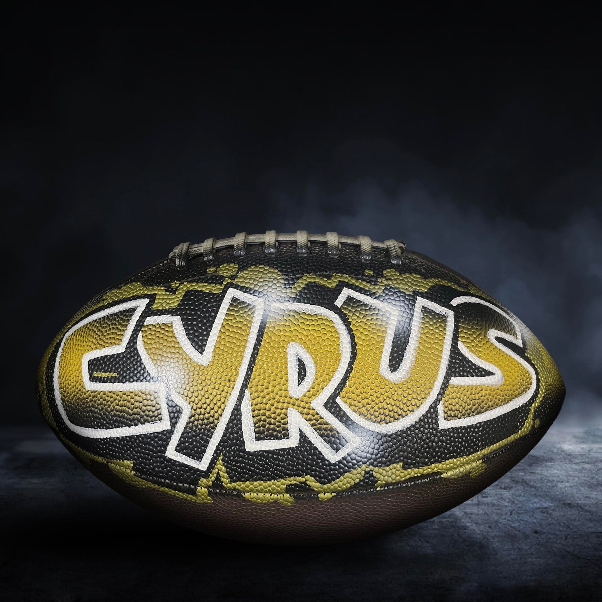 A football with the word "CYRUS" painted on it, against a dark background with smoke or fog.