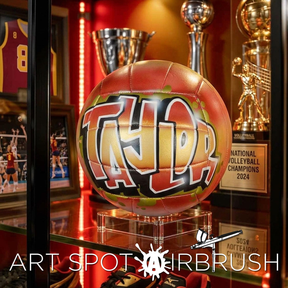 a personalized custom volleyball with the name "TAYLOR" painted on it, surrounded by various trophies and awards, including a trophy labeled "NATIONAL DOLLS BULL CHAMPIONS 2024".
