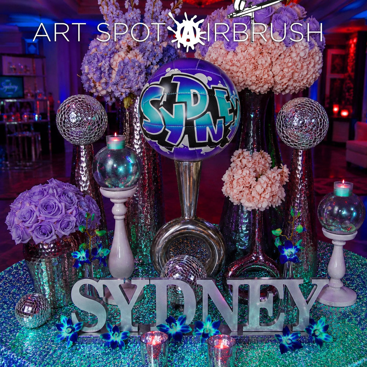 The image is a vibrant display featuring a variety of decorative elements, including a large purple balloon with the word "SYDNEY" written on it, surrounded by colorful flowers and shiny vases. The overall scene appears to be a festive or celebratory arrangement, possibly for a special event or occasion.