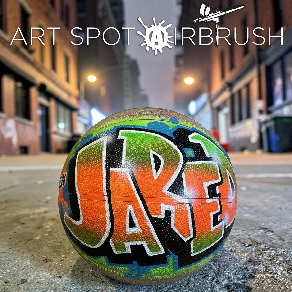 A basketball with the name "Jae" painted on it, sitting on a concrete surface in an urban setting at night.