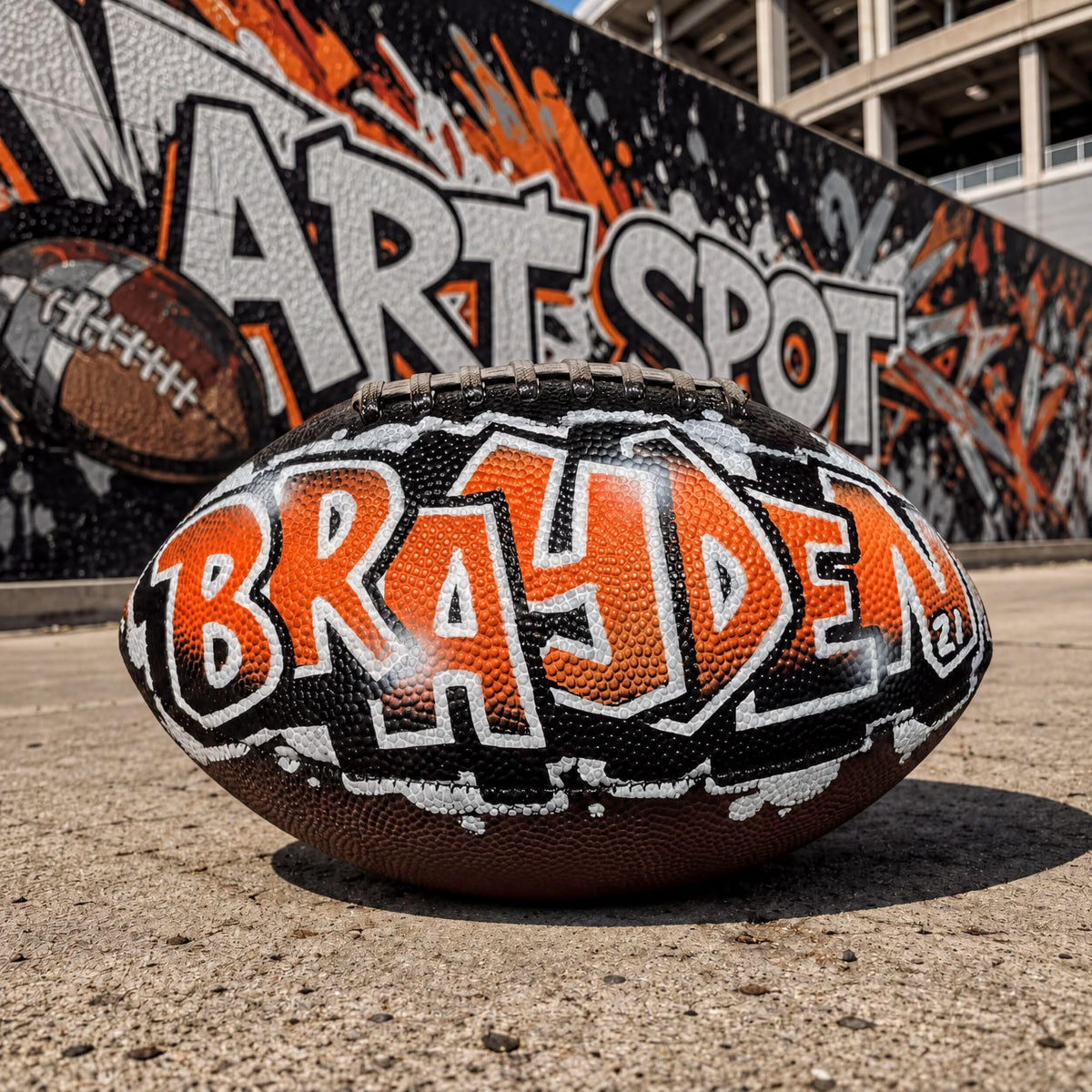 A custom football personalized with the name "Brayden" painted on it, sitting on a concrete surface in front of a graffiti-covered wall.