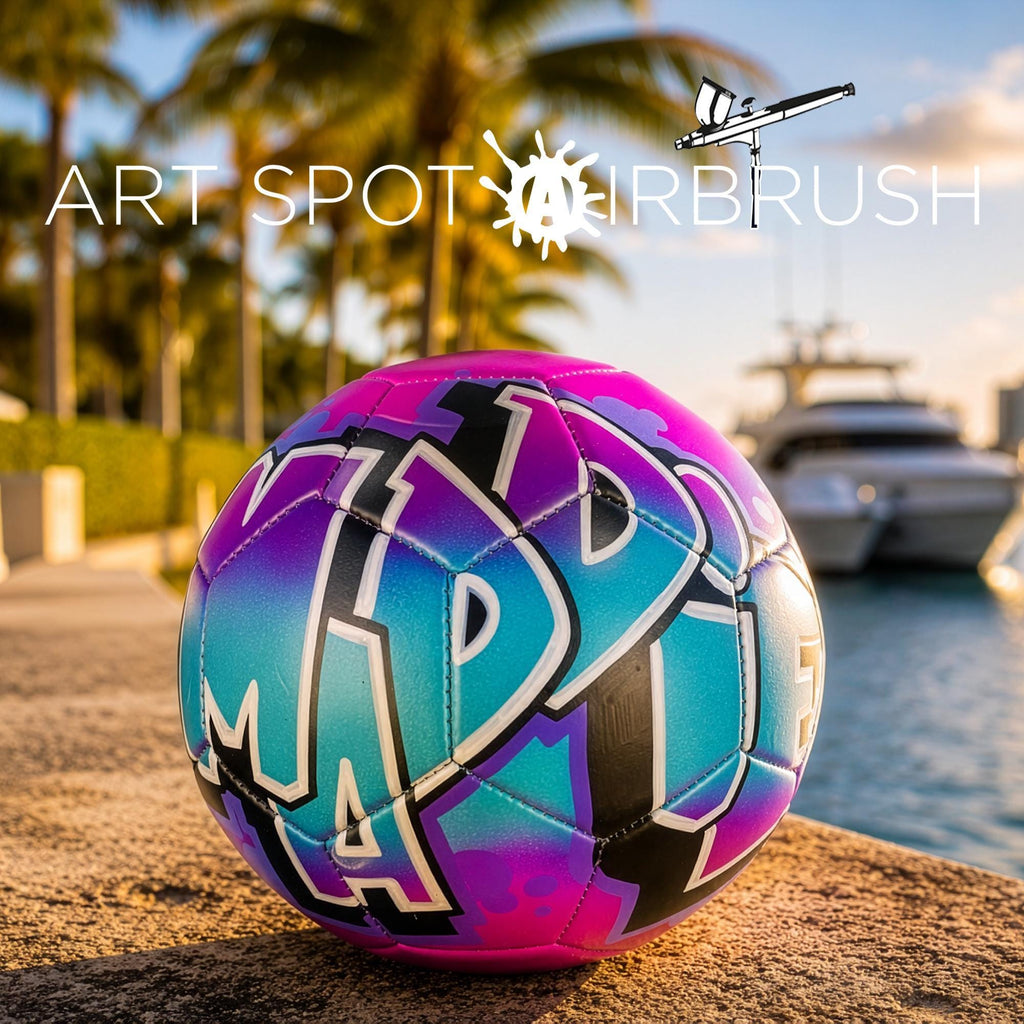 A colorful, graffiti-style soccer ball is placed on a sandy beach, with palm trees and a boat visible in the background.