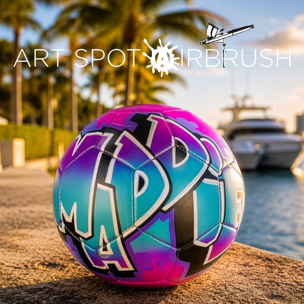 A colorful, graffiti-style soccer ball is placed on a sandy beach, with palm trees and a boat visible in the background.