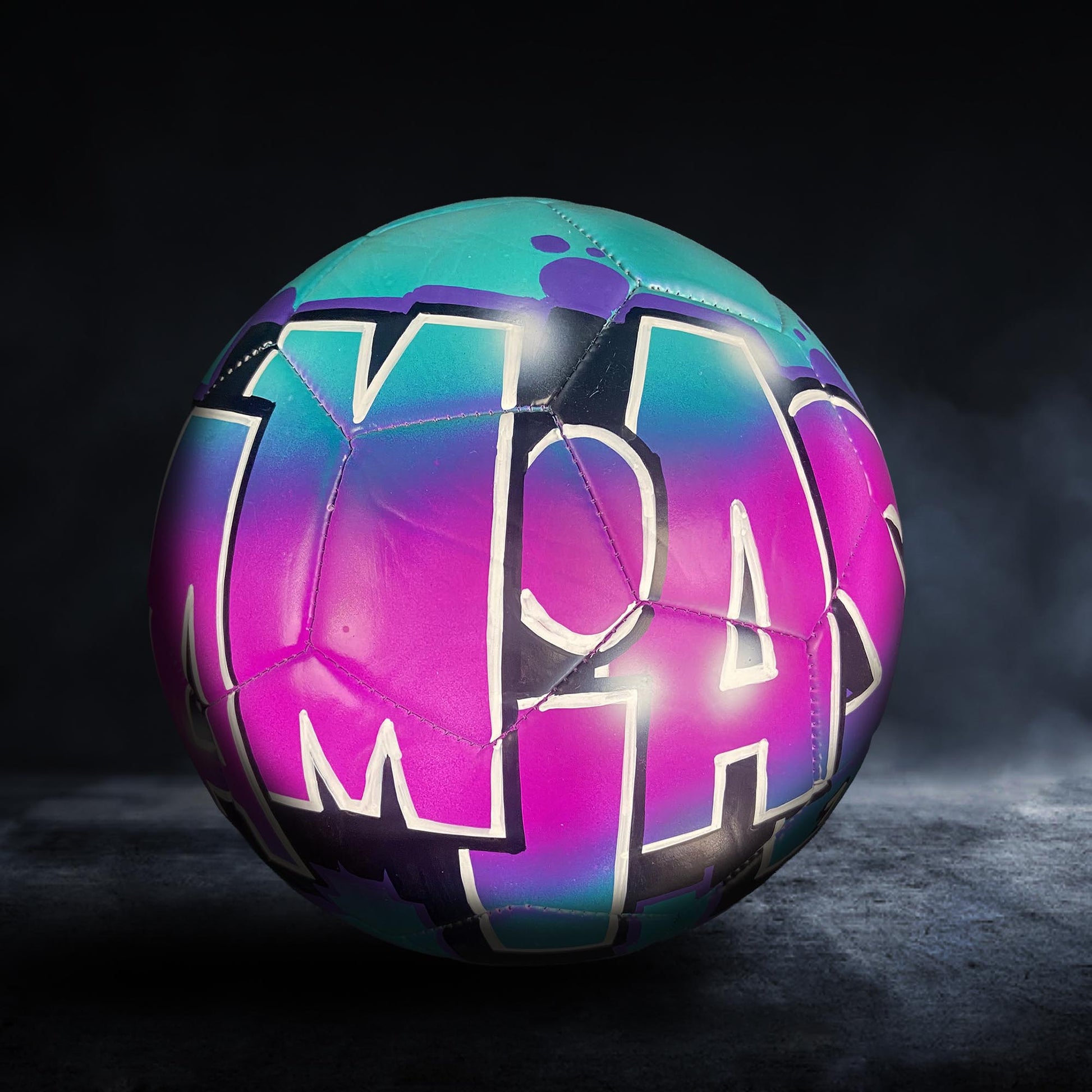 A colorful soccer ball with the word "MAY" painted on it, set against a dark background.