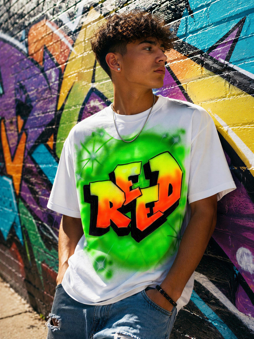 A young man wearing a white t-shirt with a colorful graffiti design stands in front of a vibrant, colorful graffiti wall.