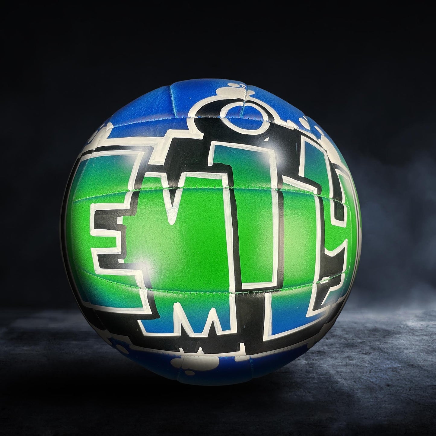 A colorful volleyball with the word "EM" painted on it, set against a dark background.