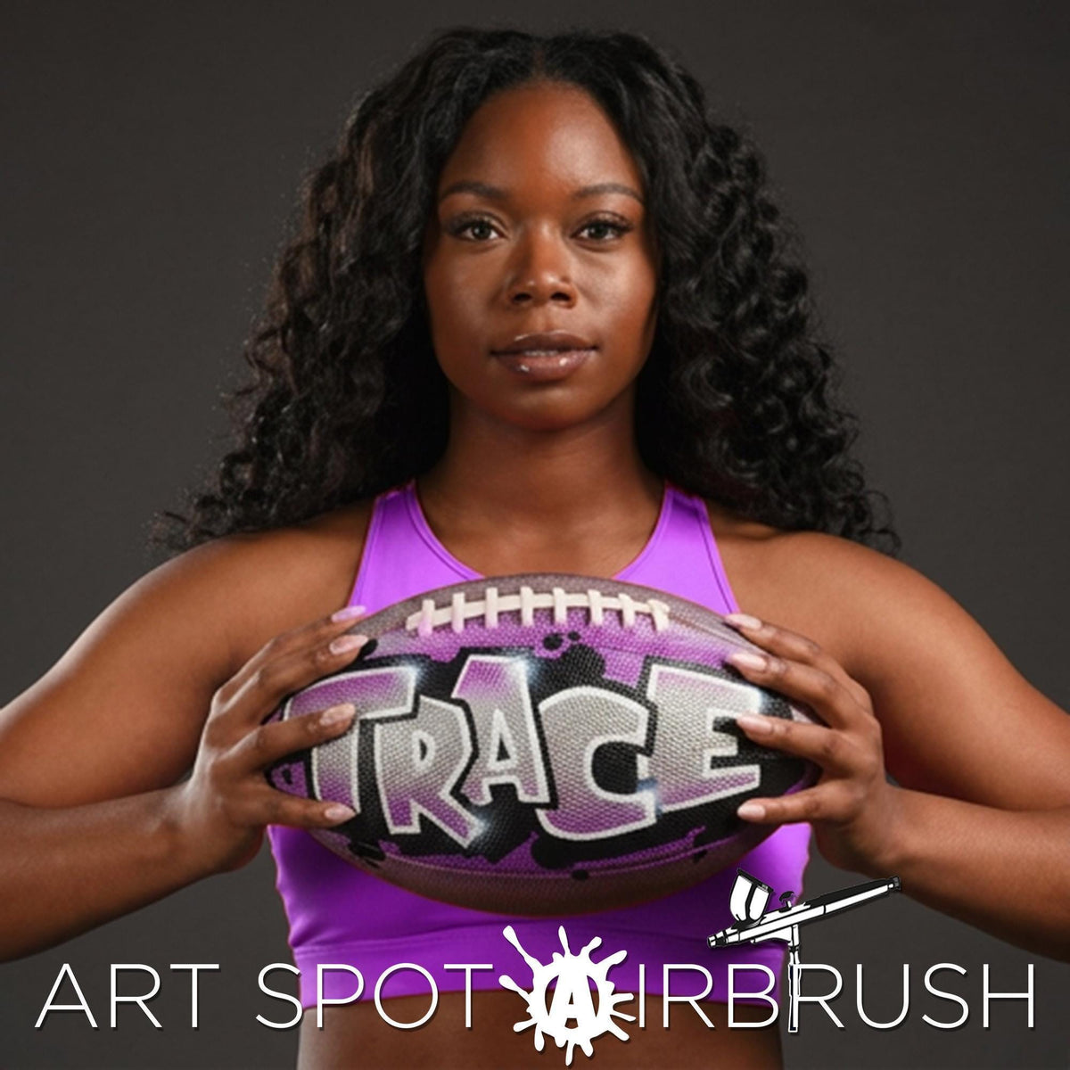 A black woman with long curly hair is holding a custom football personalized with the word "TRACE" on it.