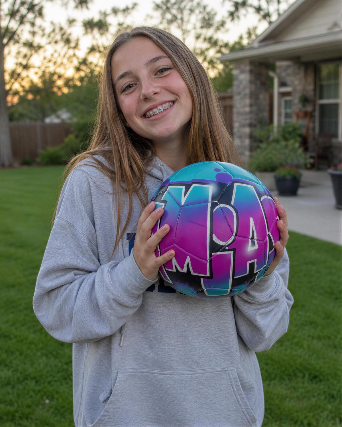 A young woman is holding a colorful soccer ball with the word "LAS" written on it, smiling and posing in front of a house.