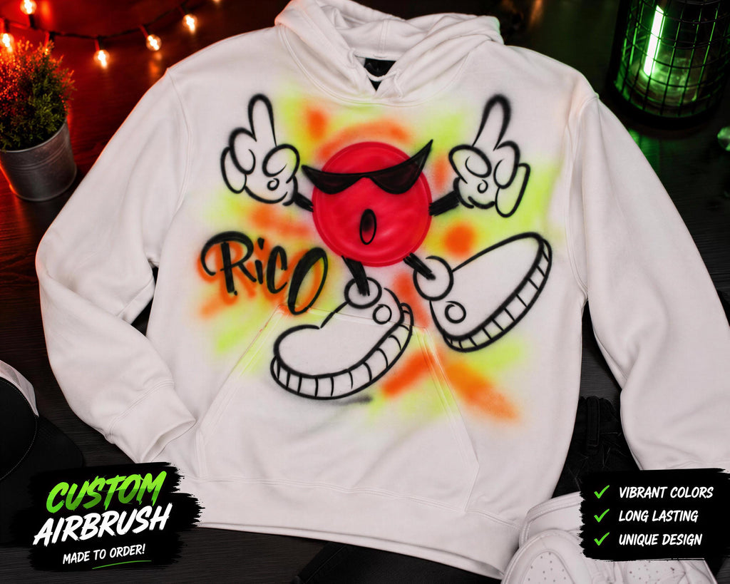 A white hoodie with a custom airbrush design featuring a red character with a face and sunglasses, along with the text "Rico" and a colorful background.