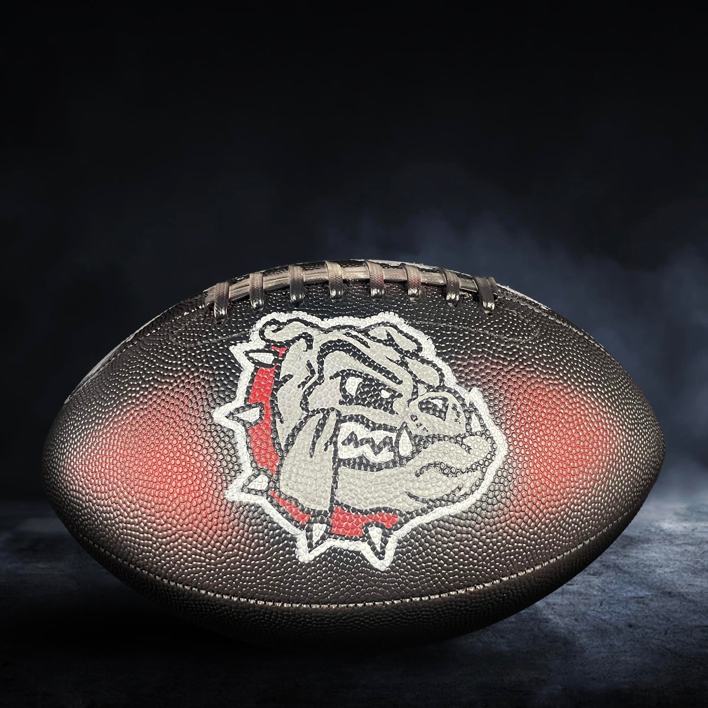 A football with a bulldog logo on it, placed on a dark surface with smoke or fog in the background.