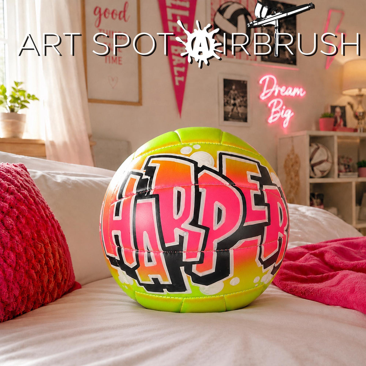 A custom volleyball with the name "HARPER" written on it, sitting on a bed with a red pillow.