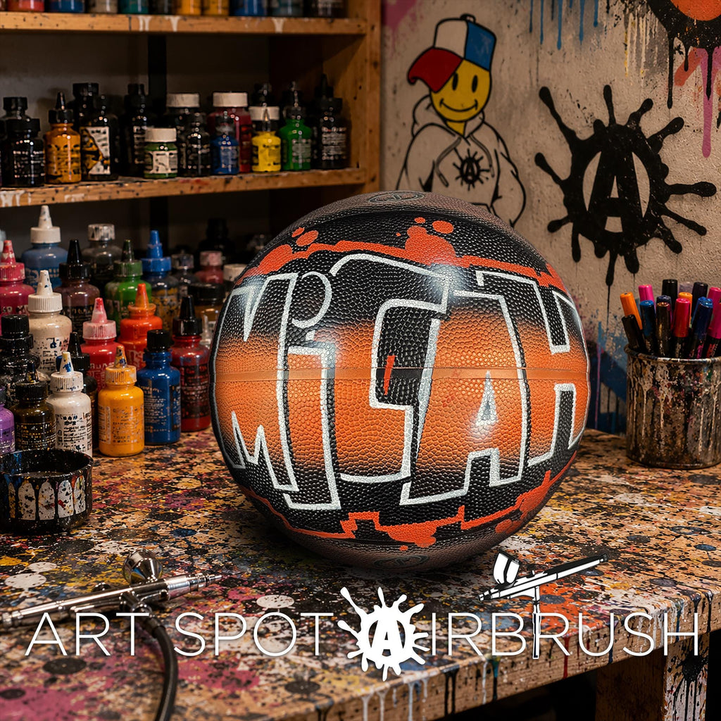A basketball with the word "MICH" painted on it, sitting on a table surrounded by various art supplies and a graffiti-style wall in the background.