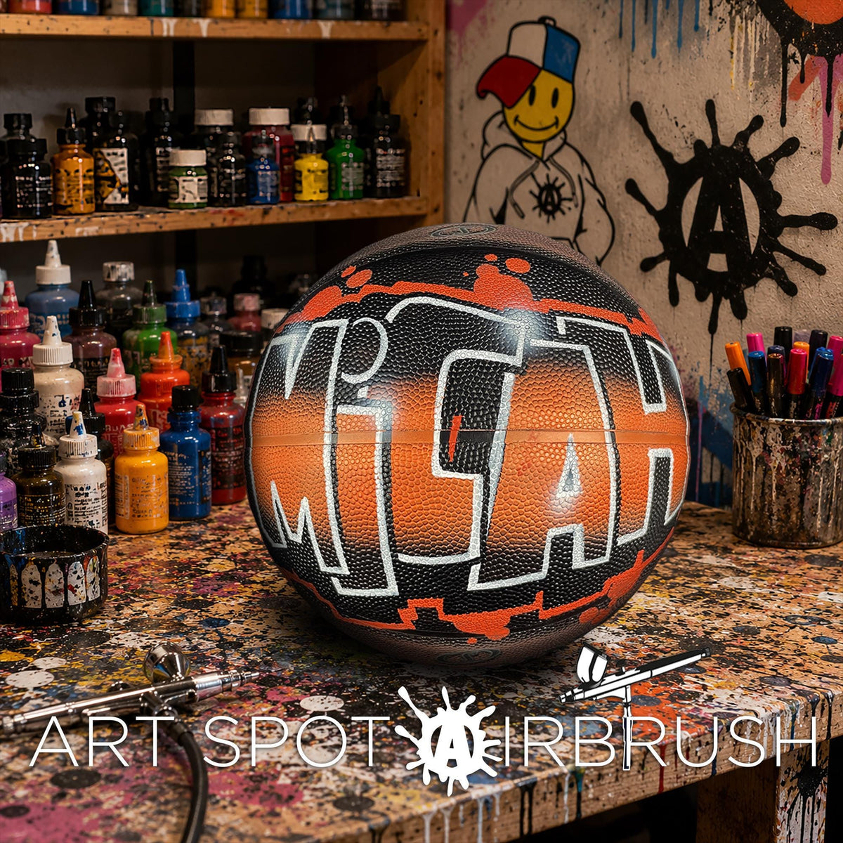 A basketball with the word "MICH" painted on it, sitting on a table surrounded by various art supplies and a graffiti-style wall in the background.