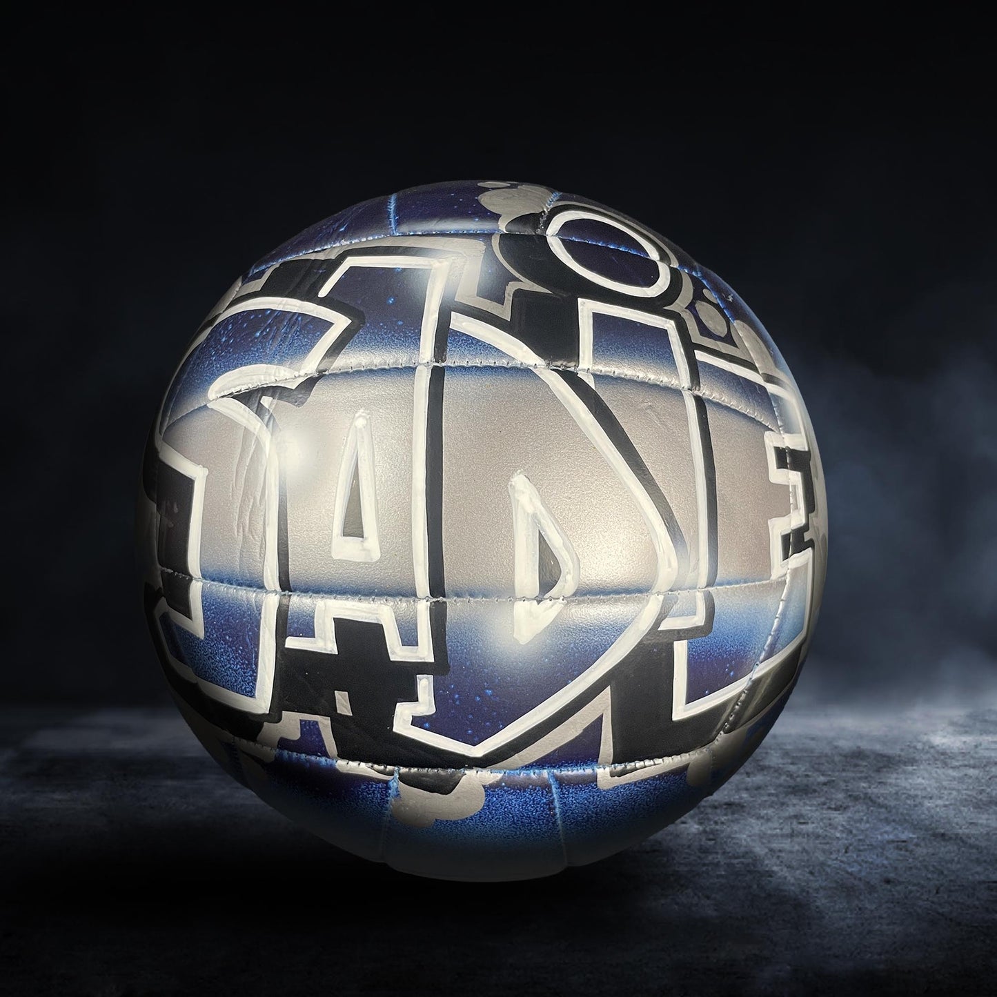 A soccer ball with a unique, graffiti-style design on it, featuring various shapes and patterns in shades of blue and gray.