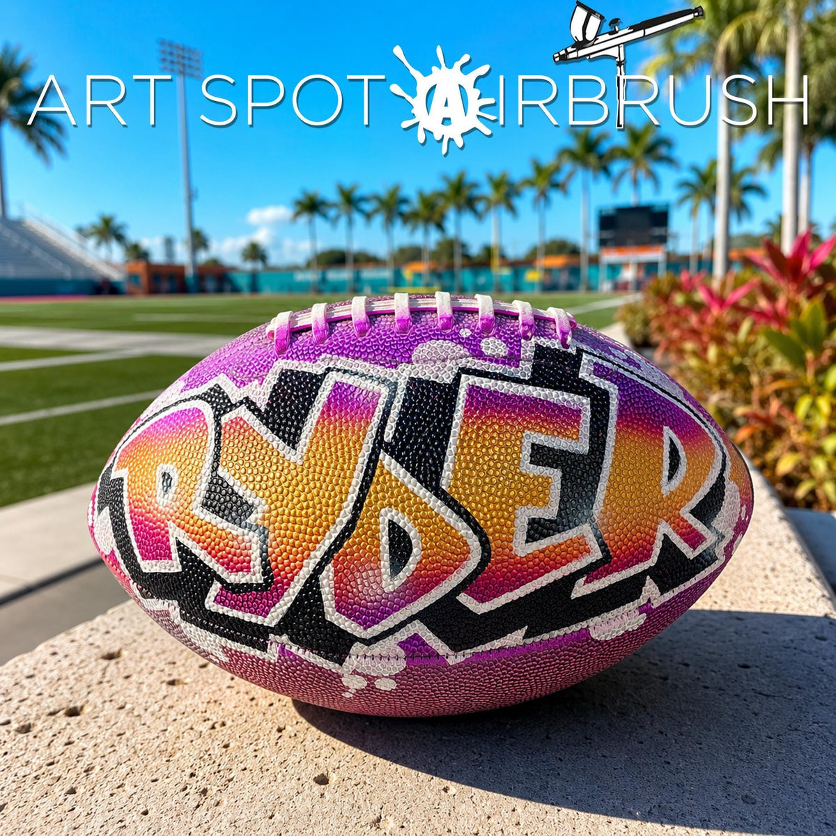A personalized custom football with the word "RYDER" painted on it, sitting on a concrete surface with palm trees and a stadium in the background.