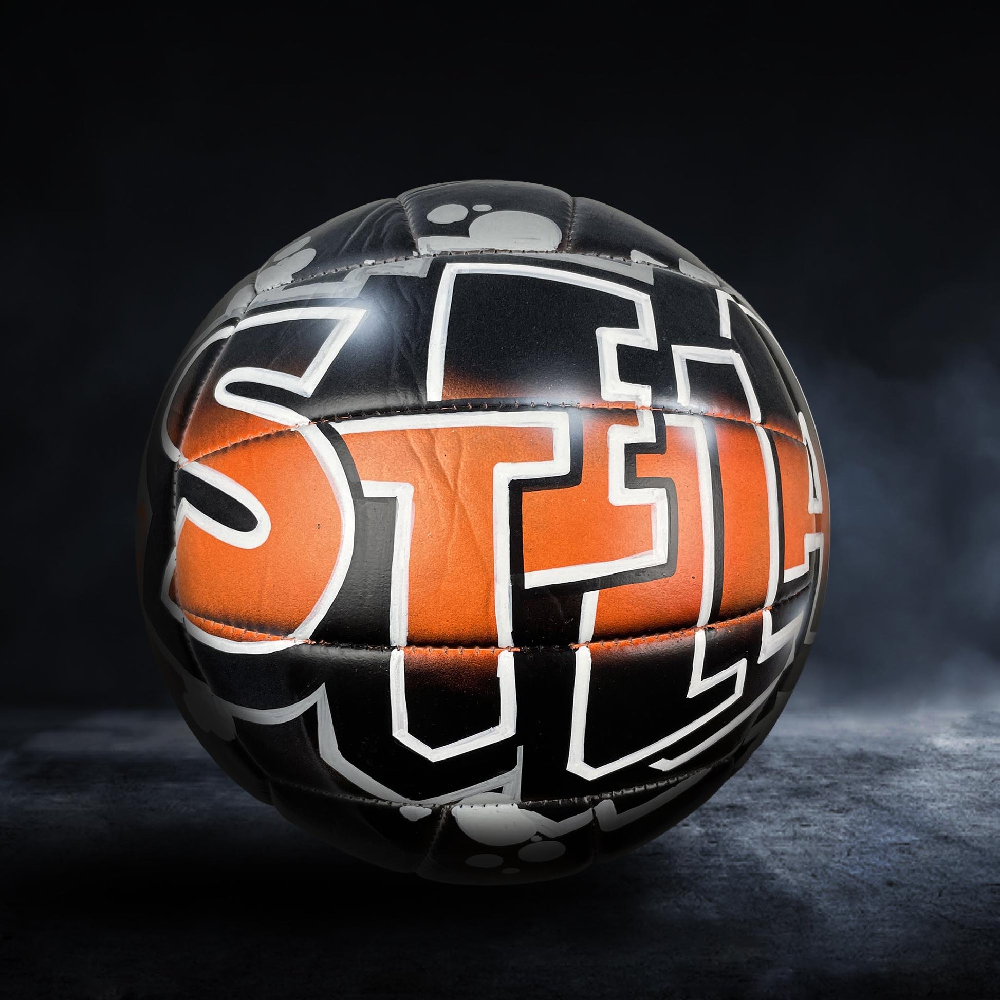 A soccer ball with the word "STEEL" painted on it, set against a dark background with smoke or fog.