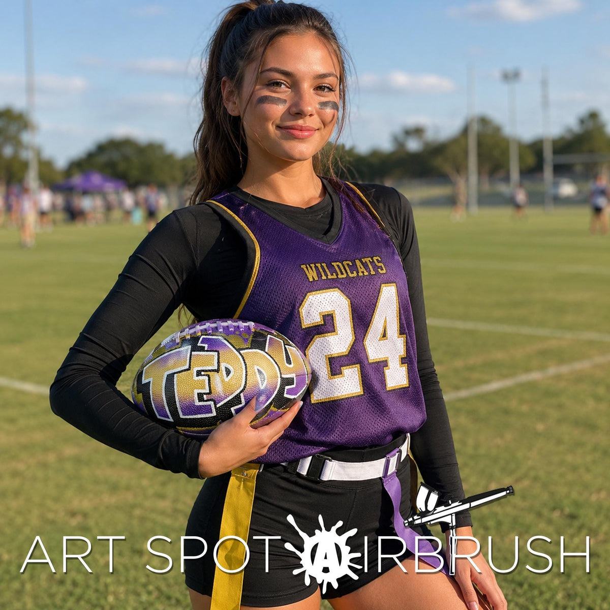 A young woman in a purple and gold sports uniform holding a purple and gold custom football personalized with the word 'Teddy' on it, standing on a grassy field.