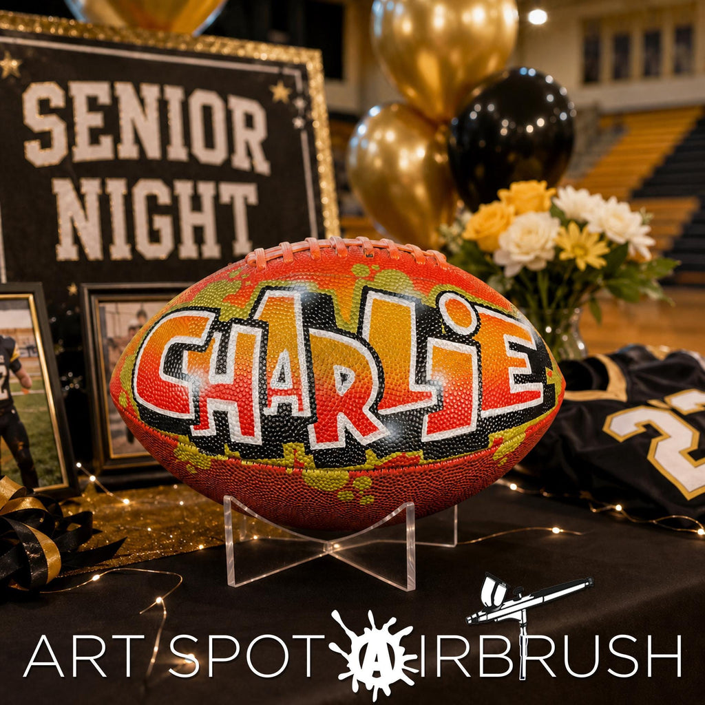 a personalized football with the name "Charlie" painted on it, displayed on a table with a sign that reads "SENIOR NIGHT" and balloons in the background.