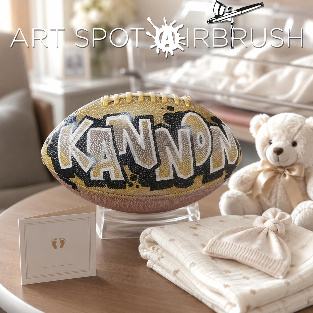 a personalized custom football with the word "KANNON" painted on it, placed on a table next to a teddy bear and some baby items.
