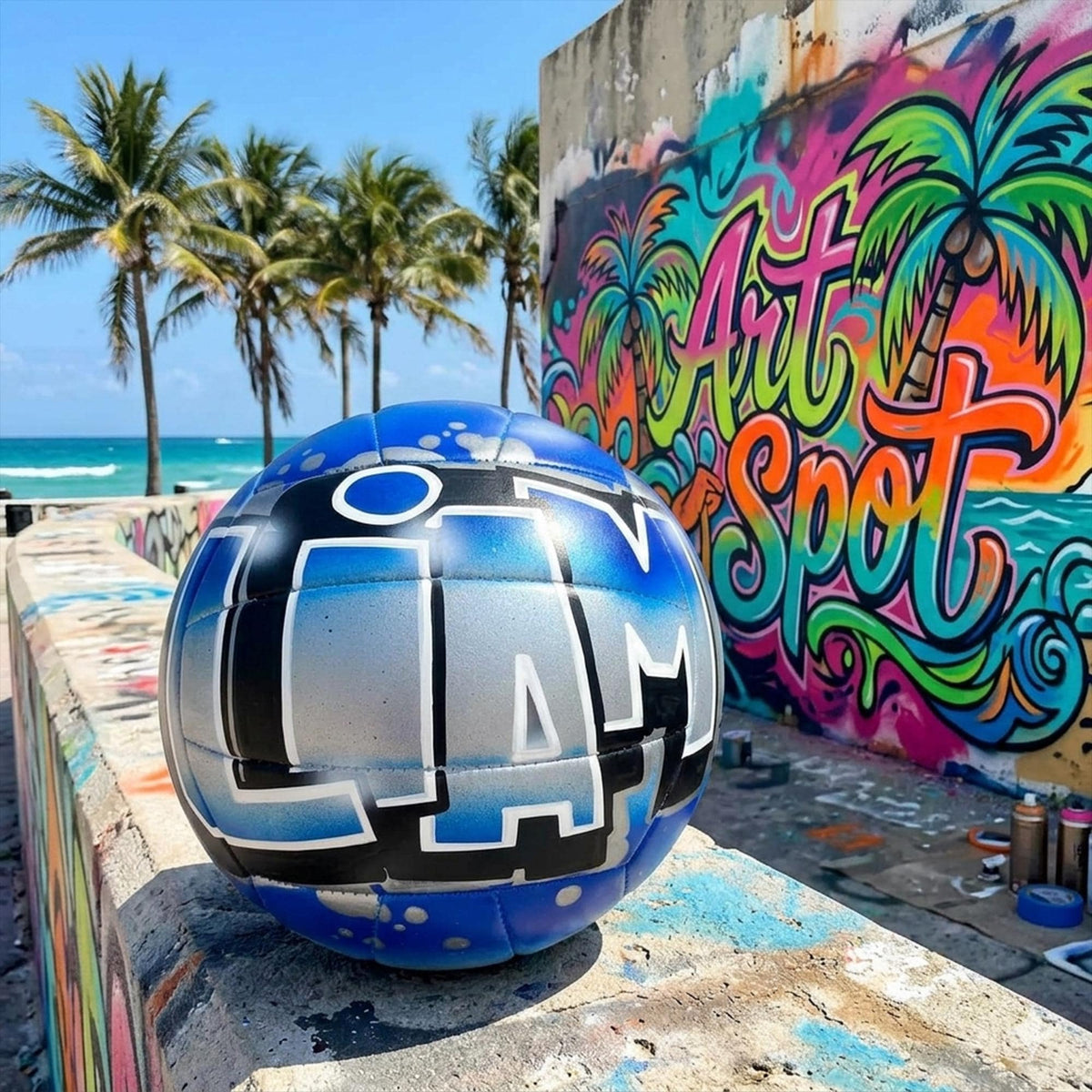 A blue and silver graffiti-style custom volleyball with the word "LIAM" painted on it, sitting on a concrete ledge in front of a colorful graffiti mural with the name ART SPOT.