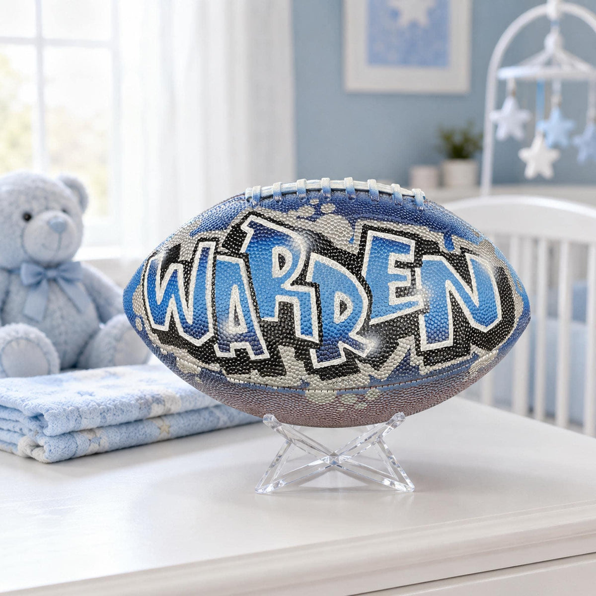 A personalized custom football with the name "WARREN" written on it, displayed on a stand in a room with a teddy bear and other baby-related items.