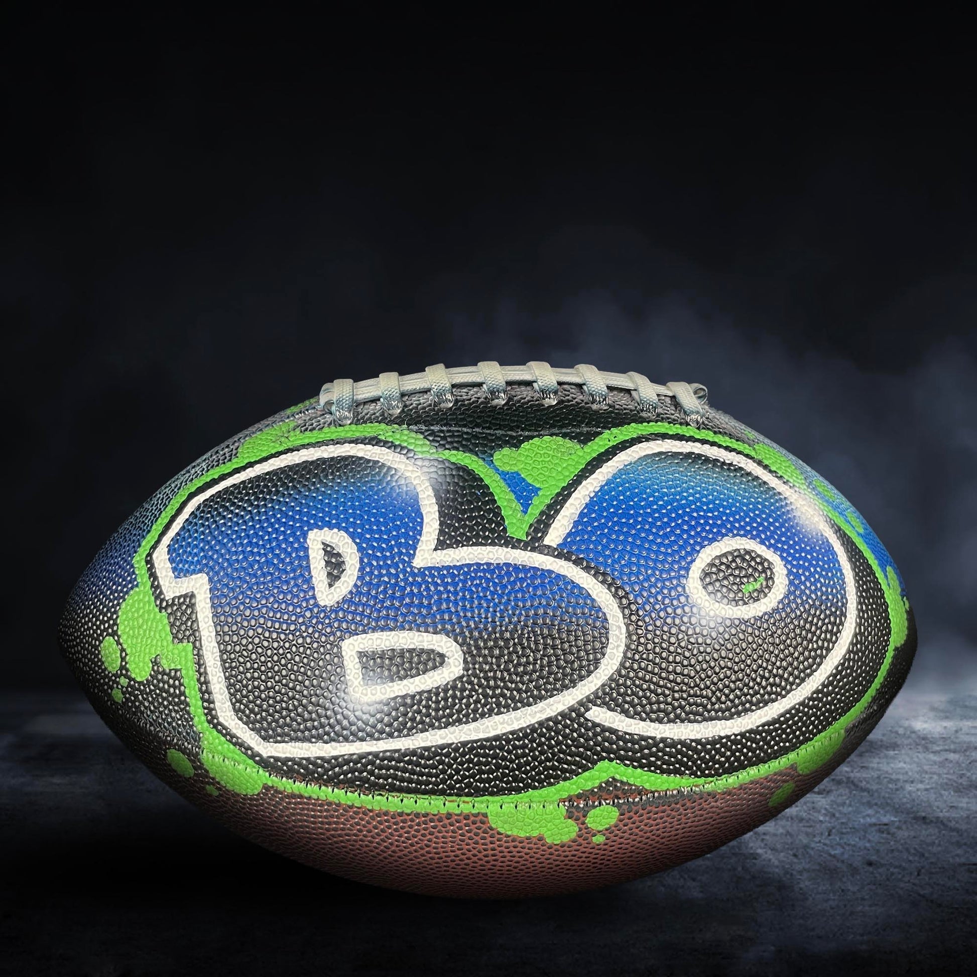A football with the word "B" painted on it, set against a dark background with smoke or fog.