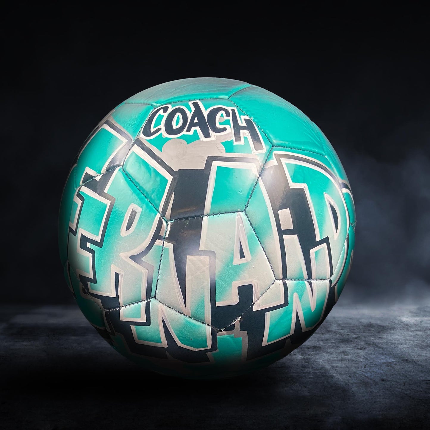 A soccer ball with the word "COACH" written on it in teal and white colors.