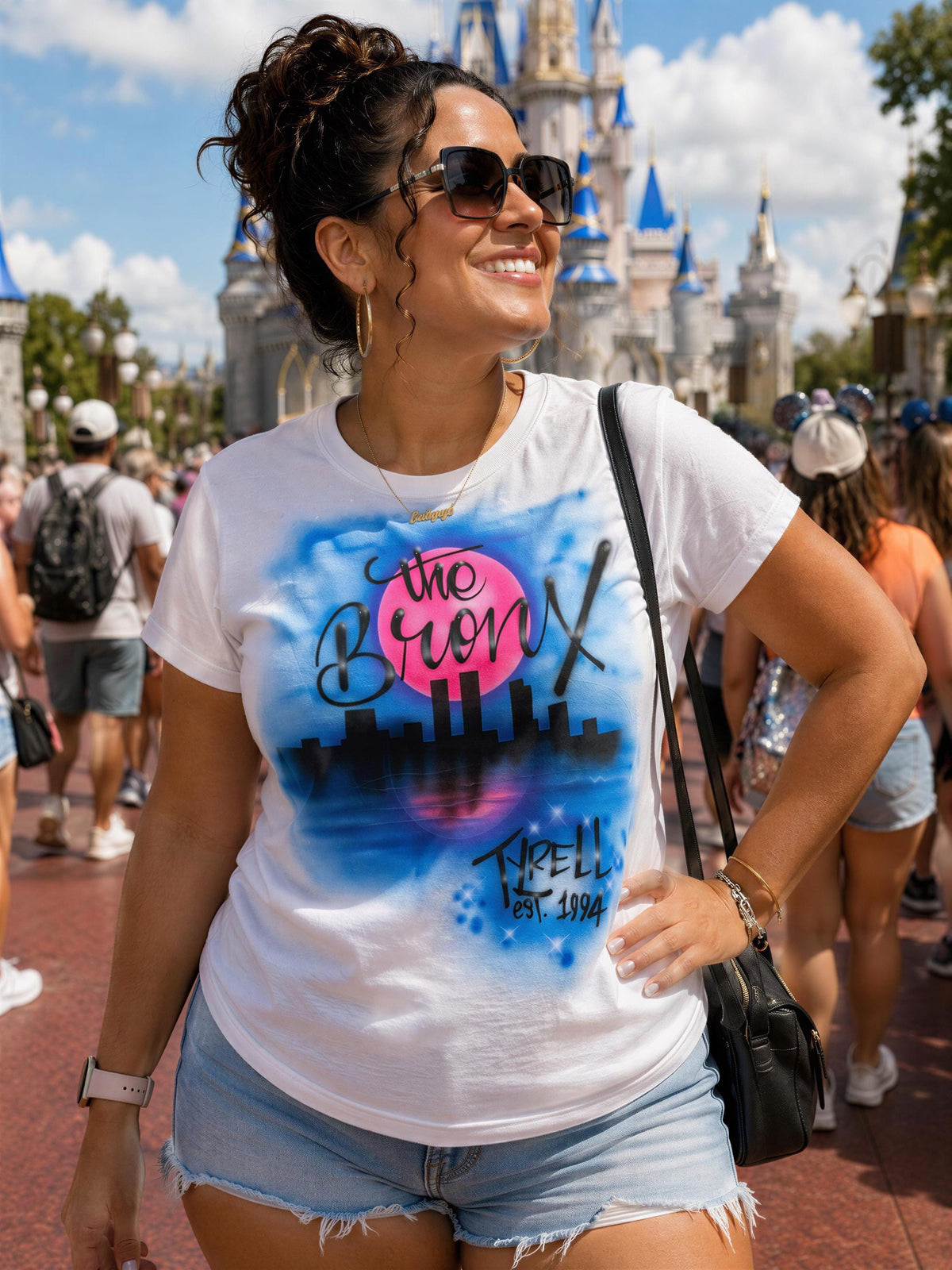 A woman wearing a white t-shirt with a city skyline design and sunglasses stands in front of a Disney World castle.