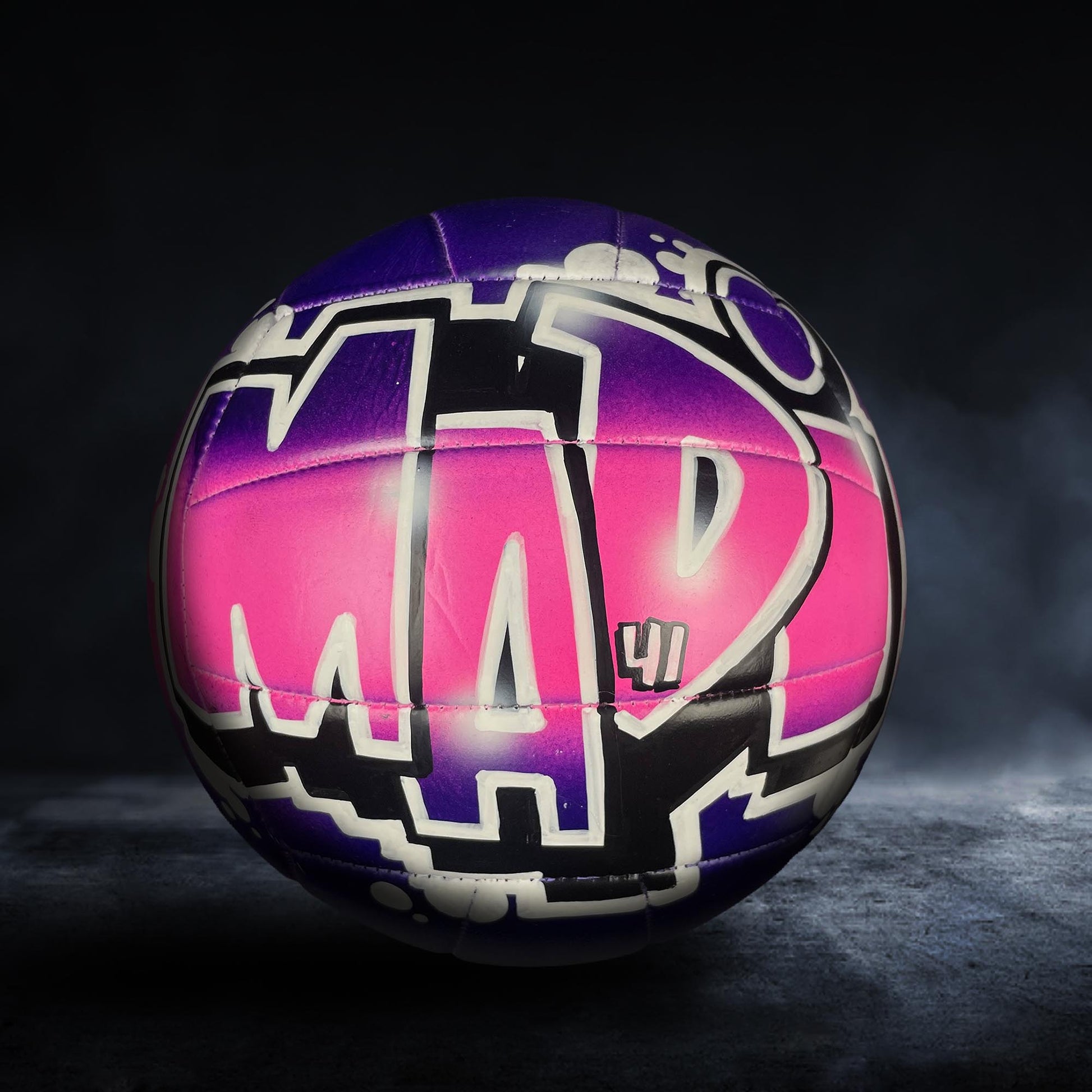 A colorful, graffiti-style volleyball with the word "M" painted on it, set against a dark background.