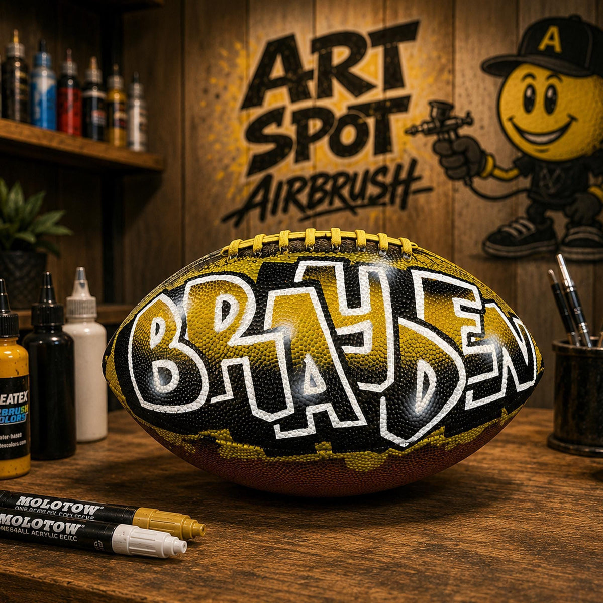 A custom football personalized with the name "BRAYDEN" painted on it, sitting on a wooden surface. In the background, there is a wall with graffiti and a cartoon character. Various art supplies, such as markers and paint, are visible on the table.