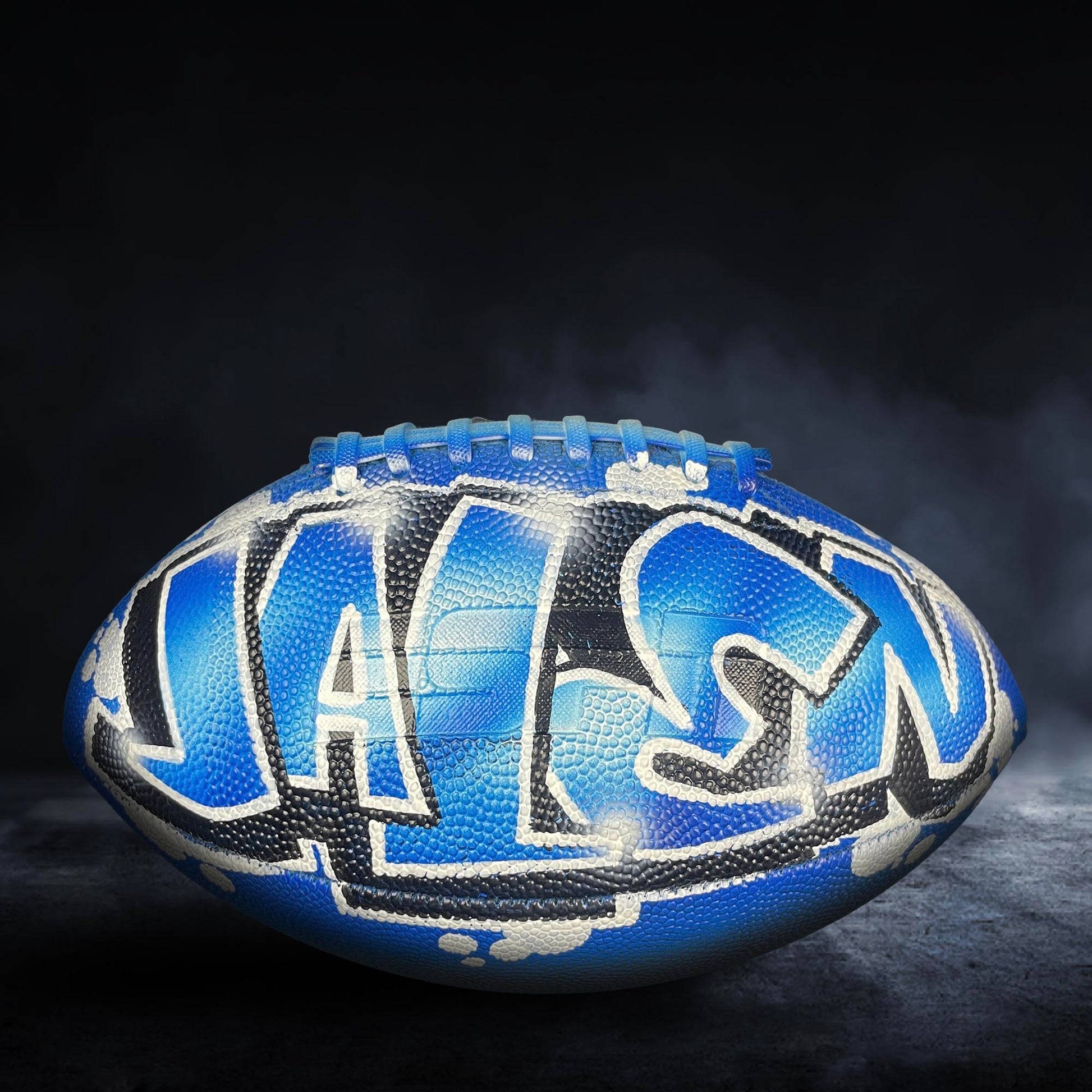 A blue and black football with graffiti on it, sitting on a dark surface with smoke in the background.