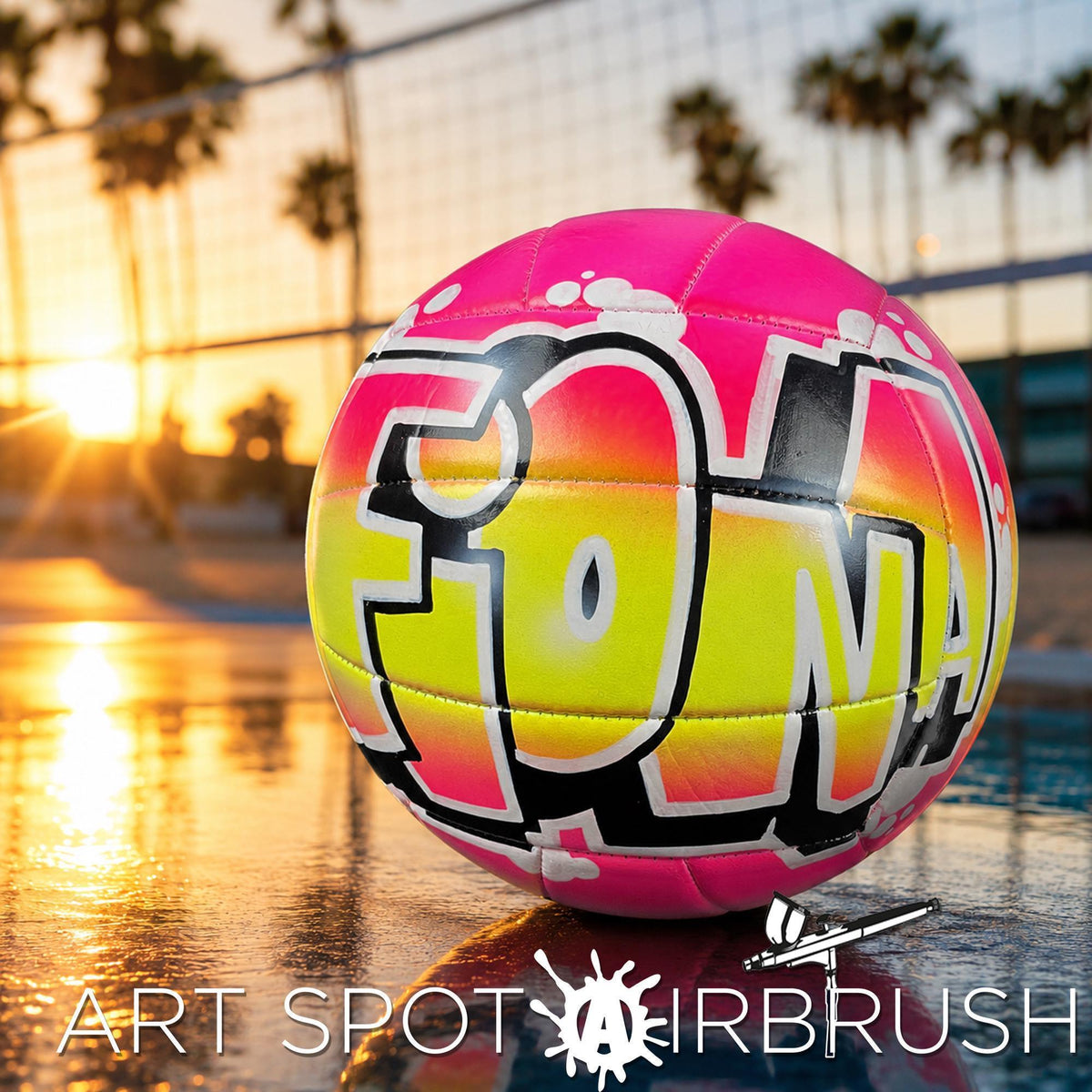 A colorful custom volleyball personalized with the word "FIONA" painted on it, sitting on a reflective surface with palm trees and a sunset in the background.