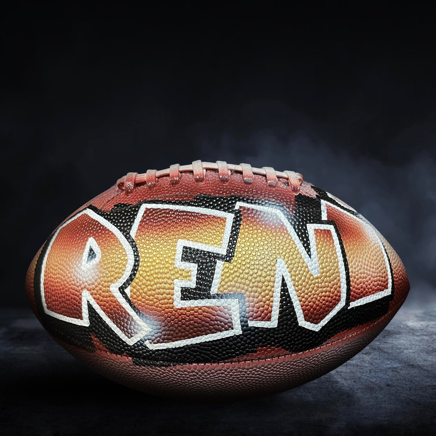 A football with the word "REN" painted on it, sitting on a dark surface with smoke or fog in the background.