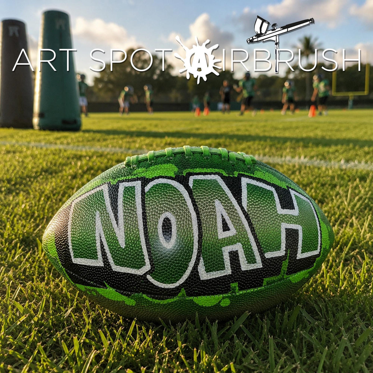 A green and black personalized custom football with the word "Noah" painted on it, sitting on a grassy field with a group of people in the background.