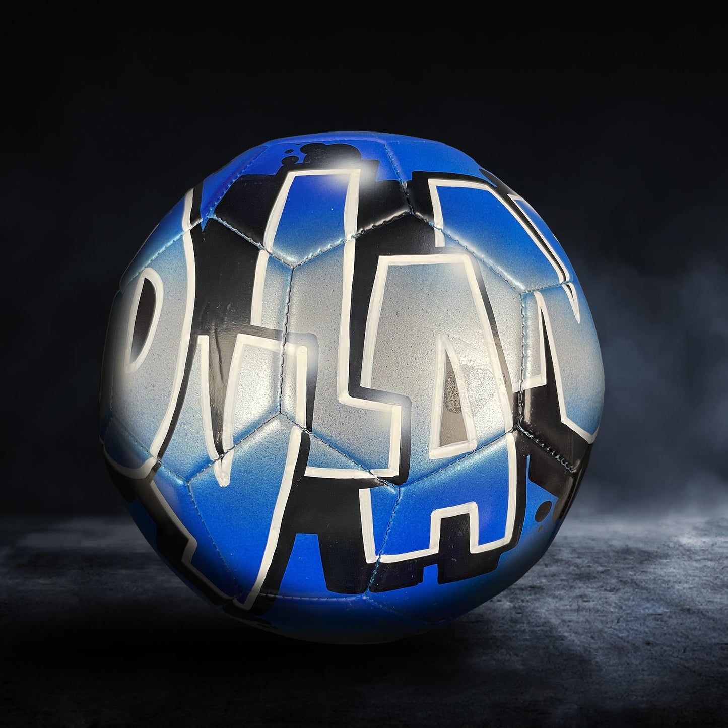 A blue and black soccer ball with the word "OLA" painted on it, sitting on a dark surface.