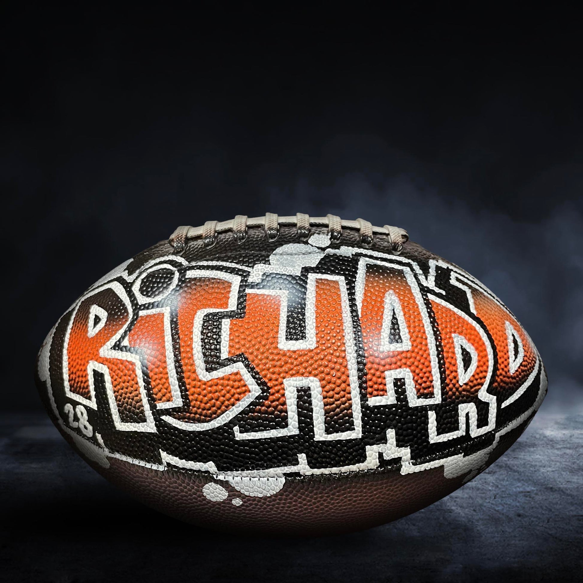 A football with the name "RICHARD" painted on it, sitting on a dark surface with a misty background.